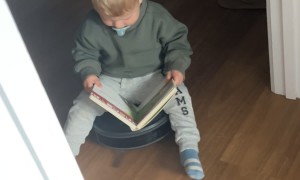 Toddler Reads a Book While Riding Robot Vacuum
